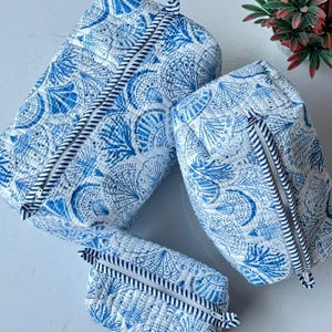 May include: Three quilted fabric pouches in varying sizes, featuring a white base with a blue seashell and coral print. Each pouch has a top zipper closure with a black and white striped fabric pull tab, showcasing a consistent quilted texture.