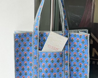 May include: A quilted blue tote bag featuring a repeating pattern of small pink flowers with green stems. The bag has two handles and a front pocket containing a white rectangular card with the text SUGAM PREMIUM SUITINGS printed on it.