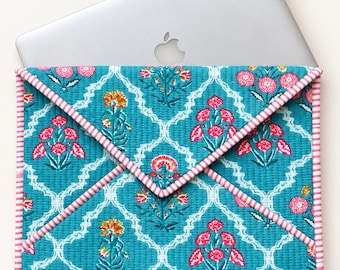 May include: A quilted laptop sleeve featuring a teal background with a white lattice pattern and pink and yellow floral motifs. The envelope-style flap and edges are trimmed with a pink and white striped fabric, housing a silver laptop.