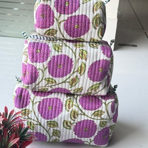 Puede incluir: Una pila de tres estuches rectangulares de tela acolchada de diferentes tama&ntilde;os. Cada estuche presenta una base blanca con un patr&oacute;n repetitivo de flores moradas y tallos verdes. Los estuches tienen un cierre de cremallera en la parte superior.