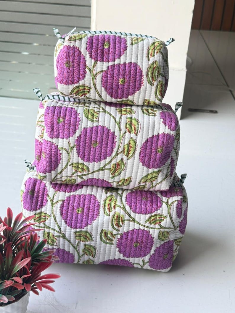 Puede incluir: Una pila de tres estuches rectangulares de tela acolchada de diferentes tama&ntilde;os. Cada estuche presenta una base blanca con un patr&oacute;n repetitivo de flores moradas y tallos verdes. Los estuches tienen un cierre de cremallera en la parte superior.
