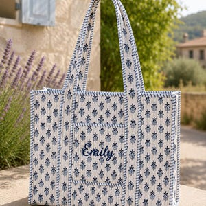 Puede incluir: Un bolso tote acolchado con base blanca y un patrón floral repetido en azul oscuro. El bolso tiene dos asas largas y un bolsillo frontal bordado con el nombre Emily en letra cursiva azul oscuro. Los bordes tienen un ribete azul.