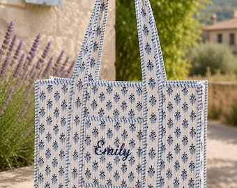 May include: A quilted tote bag featuring a white base with a repeating dark blue floral block print pattern. The bag has two long handles and a front pocket embroidered with the name Emily in dark blue script. The edges are trimmed in blue.