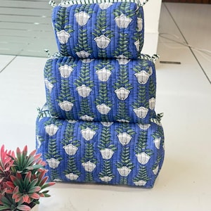 Puede incluir: Una pila de tres bolsas de almacenamiento rectangulares de tela acolchada en diferentes tama&ntilde;os. Cada bolsa presenta un fondo azul con un patr&oacute;n floral blanco repetitivo y tallos verdes. Las bolsas tienen peque&ntilde;as cintas de tela en las esquinas.