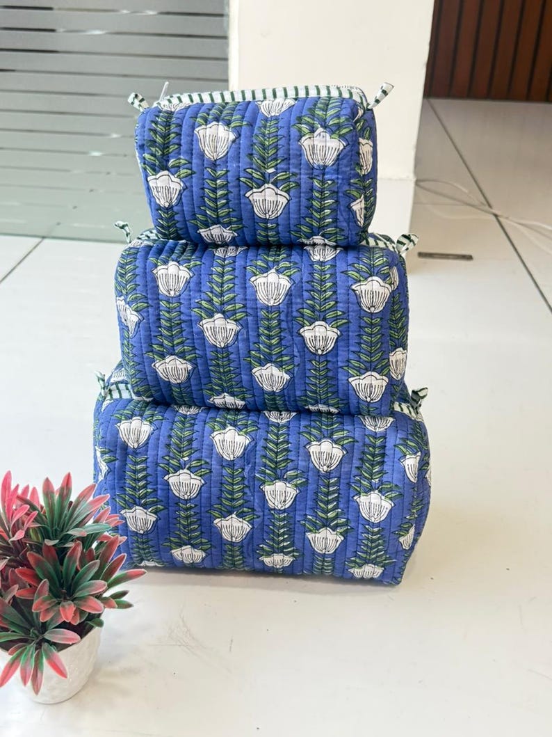 Puede incluir: Una pila de tres bolsas de almacenamiento rectangulares de tela acolchada en diferentes tama&ntilde;os. Cada bolsa presenta un fondo azul con un patr&oacute;n floral blanco repetitivo y tallos verdes. Las bolsas tienen peque&ntilde;as cintas de tela en las esquinas.