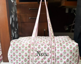 May include: A quilted duffel bag featuring a white base with a repeating pattern of small pink flowers and green stems. The bag has two long shoulder straps and a rectangular front pocket embroidered with the name Noa in black thread.