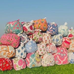May include: A large collection of quilted fabric pouches featuring various floral block print patterns in vibrant colors including red, pink, blue, orange, and green. Each rectangular pouch has a small white fabric loop on the top edge.
