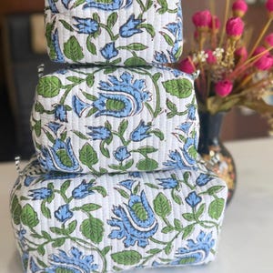 Puede incluir: Una pila de tres estuches acolchados rectangulares con una base blanca y un patr&oacute;n repetitivo de flores azules y hojas verdes. Cada estuche tiene una forma estructurada con l&iacute;neas de costura horizontales visibles en la superficie.