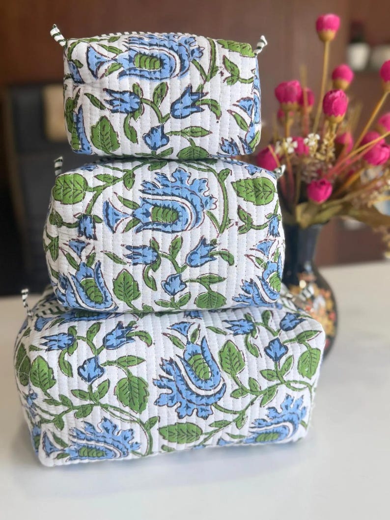 Puede incluir: Una pila de tres estuches acolchados rectangulares con una base blanca y un patr&oacute;n repetitivo de flores azules y hojas verdes. Cada estuche tiene una forma estructurada con l&iacute;neas de costura horizontales visibles en la superficie.