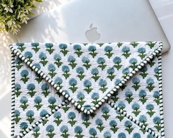 May include: A quilted laptop sleeve featuring a white base with a repeating pattern of blue flowers and green stems. The envelope-style case has a triangular flap and is finished with a green and white striped fabric trim along the edges.