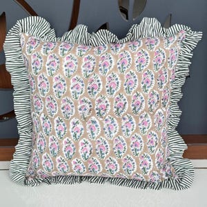 May include: Square cushion with a floral design in pink, blue, and white on a tan base. It features a ruffled border with green and white stripes. This cushion is designed for home decor.