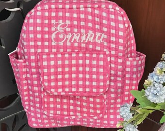 May include: A quilted backpack featuring a pink and white gingham check pattern. The front includes a rectangular pocket and the name Emma embroidered in white script lettering. The bag has side pockets and a rounded top zipper closure.