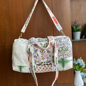 May include: A quilted duffel bag featuring a white base with a multi-colored floral and botanical print. The bag includes two short carrying handles and a long, adjustable shoulder strap, all finished with gold-toned metal hardware clips.
