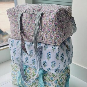 May include: A stack of three rectangular quilted duffel bags featuring floral block print patterns. The top bag is beige with pink and blue flowers, the middle is white with blue flowers, and the bottom is white with blue and green floral vines.