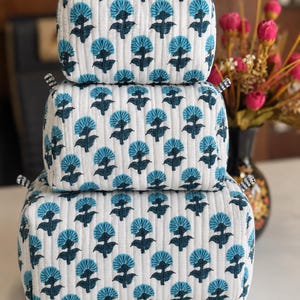 Puede incluir: Un conjunto de tres estuches acolchados rectangulares apilados verticalmente, con una base de tela blanca y un patr&oacute;n repetitivo de motivos florales en azul oscuro y azul claro. Cada estuche tiene un borde a cuadros blanco y negro.