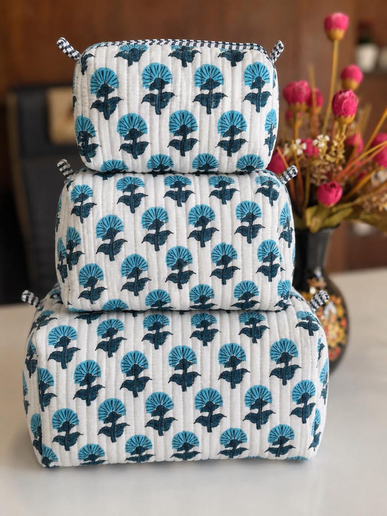 Puede incluir: Un conjunto de tres estuches acolchados rectangulares apilados verticalmente, con una base de tela blanca y un patr&oacute;n repetitivo de motivos florales en azul oscuro y azul claro. Cada estuche tiene un borde a cuadros blanco y negro.