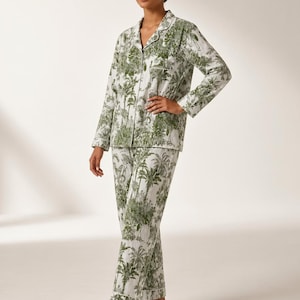 May include: A two-piece pajama set featuring a long-sleeved button-down shirt with a notched collar and matching straight-leg trousers. The fabric is white with a detailed green toile print depicting tropical palm trees and foliage patterns.