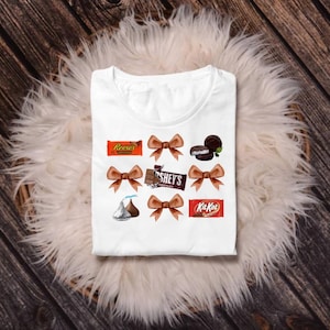 May include: White t-shirt featuring a design of various candies and brown bows. The candy brands include Reese's, Hershey's, Kit Kat, and an Oreo cookie. The shirt is laid on a white fluffy surface against a wooden background.