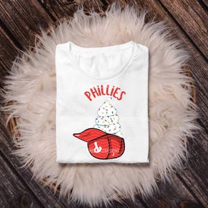 May include: White t-shirt featuring the word "Phillies" in red script above a whimsical design. The design includes a baseball cap with a soft-serve ice cream cone, decorated with colorful sprinkles. The shirt is folded on a fluffy, white surface.