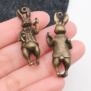 May include: Two antique gold-toned rabbit charms with a detailed design. The rabbit on the left is holding a pocket watch and the rabbit on the right is holding a flower.
