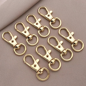 May include: A set of ten gold-colored metal keychains with a spring-loaded clasp. The keychains are all the same size and shape, and they are arranged in a circle on a light brown surface.