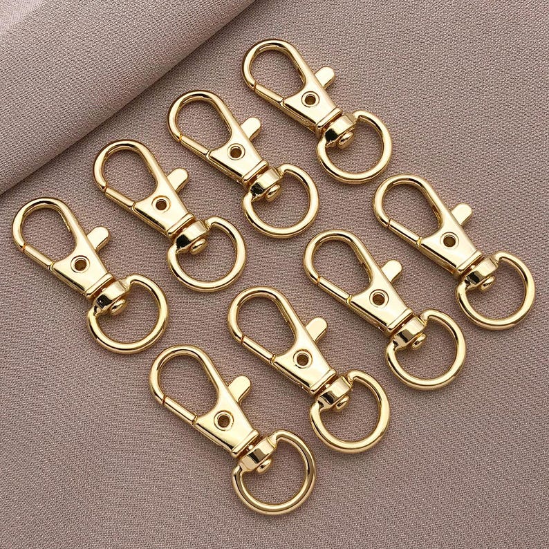 May include: A set of ten gold-colored metal keychains with a spring-loaded clasp. The keychains are all the same size and shape, and they are arranged in a circle on a light brown surface.