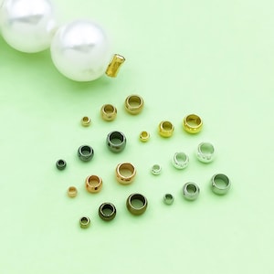 May include: An assortment of metal beads in various sizes and colors, including gold, silver, bronze, and dark gray. The beads are scattered on a light green surface, with a large white pearl necklace in the upper left corner.