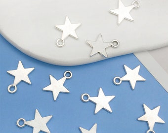 Puede incluir: Diez abalorios de estrella plateados con un bucle en la parte superior. Los abalorios están esparcidos sobre una superficie blanca y azul.