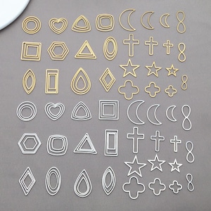 Könnte beinhalten: Eine Sammlung gold- und silberfarbener Metallanhänger in verschiedenen Formen, darunter Kreise, Herzen, Dreiecke, Quadrate, Sechsecke, Ovale, Tropfen, Rauten, Monde, Kreuze, Sterne, Kleeblätter und Unendlichkeitszeichen. Die Anhänger sind auf einer grauen Oberfläche angeordnet.