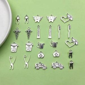 May include: A collection of silver-toned metal charms with dental and medical themes. The charms include toothbrushes, toothpaste tubes, teeth, floss containers, a medical cross, a heart, and a dental ambulance.