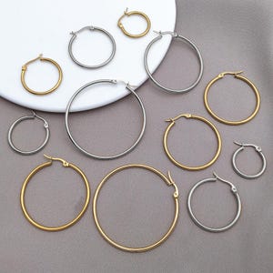 Puede incluir: Un surtido de pendientes de aro dorados y plateados de varios tamaños se colocan sobre una superficie blanca y un fondo gris. Los pendientes son de metal y tienen un diseño sencillo y clásico. Los aros varían de pequeños a grandes.