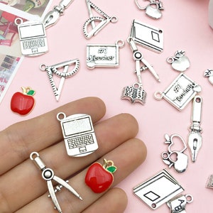 May include: Assortment of silver charm pendants with school and teacher themes. Charms include an apple, a book, a laptop, a protractor, a compass, a ruler, and a notebook. Some charms have enamel details, such as red apples and a blue notebook.