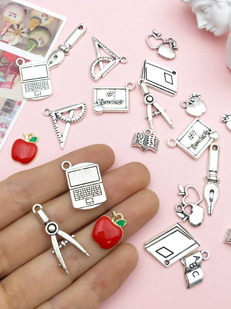 May include: Assortment of silver charm pendants with school and teacher themes. Charms include an apple, a book, a laptop, a protractor, a compass, a ruler, and a notebook. Some charms have enamel details, such as red apples and a blue notebook.