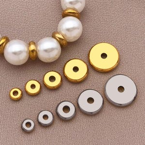 以下が含まれることがあります： 金と銀色の金属製ジュエリーパーツの詰め合わせ。中央に穴の開いた丸いスペーサービーズが含まれています。背景には、パールとゴールドビーズのブレスレットが部分的に見えます。これらのコンポーネントは、ジュエリー作りに使用されます。