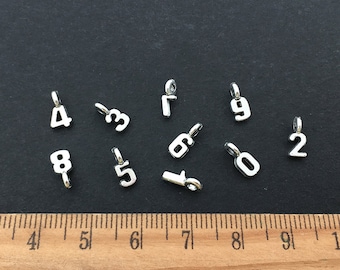 May include: Ten silver metal number charms, each with a different number from 0 to 9. The charms are arranged on a black surface with a wooden ruler in the bottom of the image.