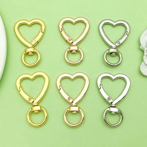 Könnte beinhalten: Sechs herzförmige Drehverschlüsse in Gold- und Silbertönen. Die Verschlüsse haben einen federbelasteten Mechanismus und einen kleinen Ring zum Befestigen. Der Hintergrund ist hellgrün.