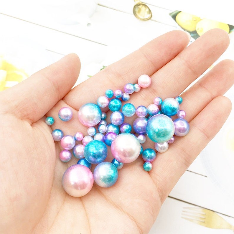 May include: A collection of iridescent pastel blue and pink pearl beads in various sizes. The beads have a pearlescent finish and are scattered in a hand.