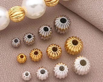 Puede incluir: Una variedad de cuentas de joyería en tonos dorados y plateados. Las cuentas incluyen cuentas doradas acanaladas, grandes cuentas de perlas blancas y pequeñas cuentas en forma de flor en oro y plata. Las cuentas están dispuestas sobre una superficie texturizada de color neutro.