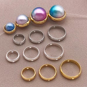 Op de afbeelding: Een assortiment van sieradenonderdelen, waaronder ronde ringen in zilver- en goudtinten, en parelachtige kralen in blauwe, paarse en turquoise tinten, elk gezet in een goudkleurige bezel. De onderdelen zijn gerangschikt op een neutraal getinte ondergrond.