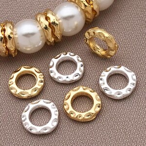Pu&ograve; includere: Primo piano di perline per gioielli color oro e argento. Le perline hanno un design circolare strutturato. Alcune perline sono disposte con grandi perle bianche e anelli strutturati color oro. Lo sfondo &egrave; un tessuto strutturato neutro.