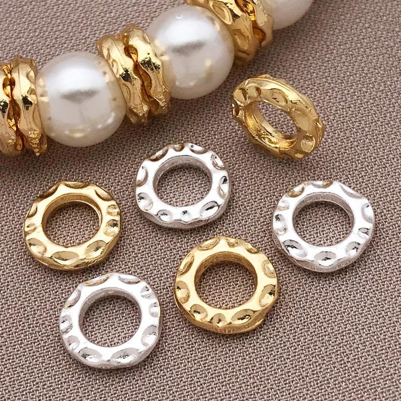 Pu&ograve; includere: Primo piano di perline per gioielli color oro e argento. Le perline hanno un design circolare strutturato. Alcune perline sono disposte con grandi perle bianche e anelli strutturati color oro. Lo sfondo &egrave; un tessuto strutturato neutro.