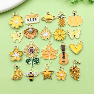 Op de afbeelding: Assortiment van 20 goudkleurige metalen bedels met emaille accenten. De bedels hebben verschillende ontwerpen, waaronder een zonnebloem, een gitaar, een ananas, een vlinder, een kat, een banaan, een bij, een ster, een blad, een hart, een bloem, een piano, een planeet, een pompoen en een schelp.