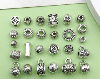 May include: A collection of 20 silver-toned metal beads with various designs, including geometric shapes, flowers, and swirls. The beads are arranged in rows on a green surface.