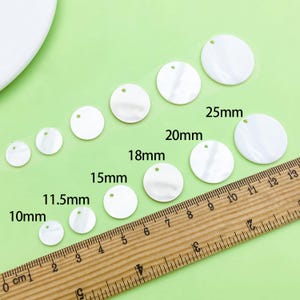 Op de afbeelding: Witte, ronde parelmoeren kralen in verschillende maten, variërend van 1 cm tot 2,5 cm, met een klein gaatje aan de bovenkant om te rijgen. Een houten liniaal is naast de kralen geplaatst voor maatvergelijking. De kralen liggen op een lichtgroen oppervlak.
