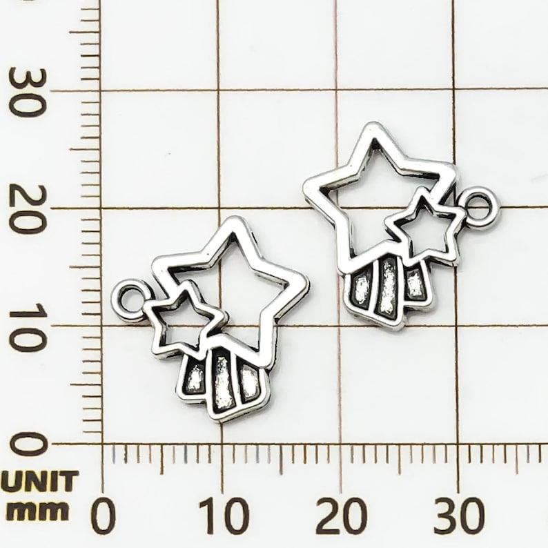 K&ouml;nnte beinhalten: Zwei silberfarbene Stern-Anh&auml;nger. Jeder Anh&auml;nger hat eine gro&szlig;e, offene Sternform mit einem kleineren Stern und einem vertikalen Balkendesign. Die Anh&auml;nger haben eine kleine Schlaufe zum Befestigen.