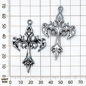 May include: Two silver-colored, ornate cross pendants. Each cross features a detailed, stylized design with pointed ends and a loop at the top for hanging. The crosses are displayed against a grid background with millimeter measurements.