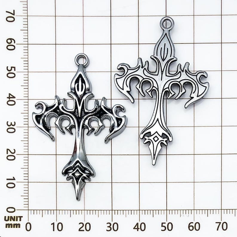 May include: Two silver-colored, ornate cross pendants. Each cross features a detailed, stylized design with pointed ends and a loop at the top for hanging. The crosses are displayed against a grid background with millimeter measurements.