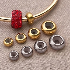 Puede incluir: Una colección de cuentas de joyería en tonos dorados y plateados. Una cuenta roja con incrustaciones de cristal está unida a una pulsera dorada. Las cuentas son redondas con un agujero en el centro. Las cuentas son para hacer joyas.