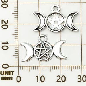 K&ouml;nnte beinhalten: Zwei silberfarbene Anh&auml;nger mit einem dreifachen Mond-Design mit einem Pentagramm in der Mitte. Die Anh&auml;nger sind etwa 2,5 cm breit. Der Hintergrund ist ein Raster mit Millimeter-Messungen.