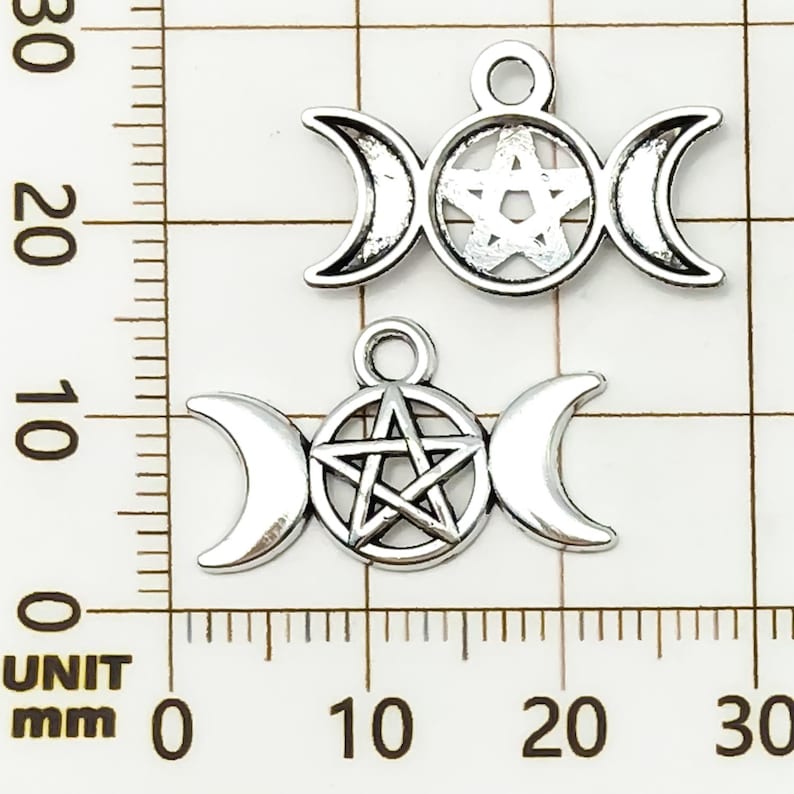 K&ouml;nnte beinhalten: Zwei silberfarbene Anh&auml;nger mit einem dreifachen Mond-Design mit einem Pentagramm in der Mitte. Die Anh&auml;nger sind etwa 2,5 cm breit. Der Hintergrund ist ein Raster mit Millimeter-Messungen.